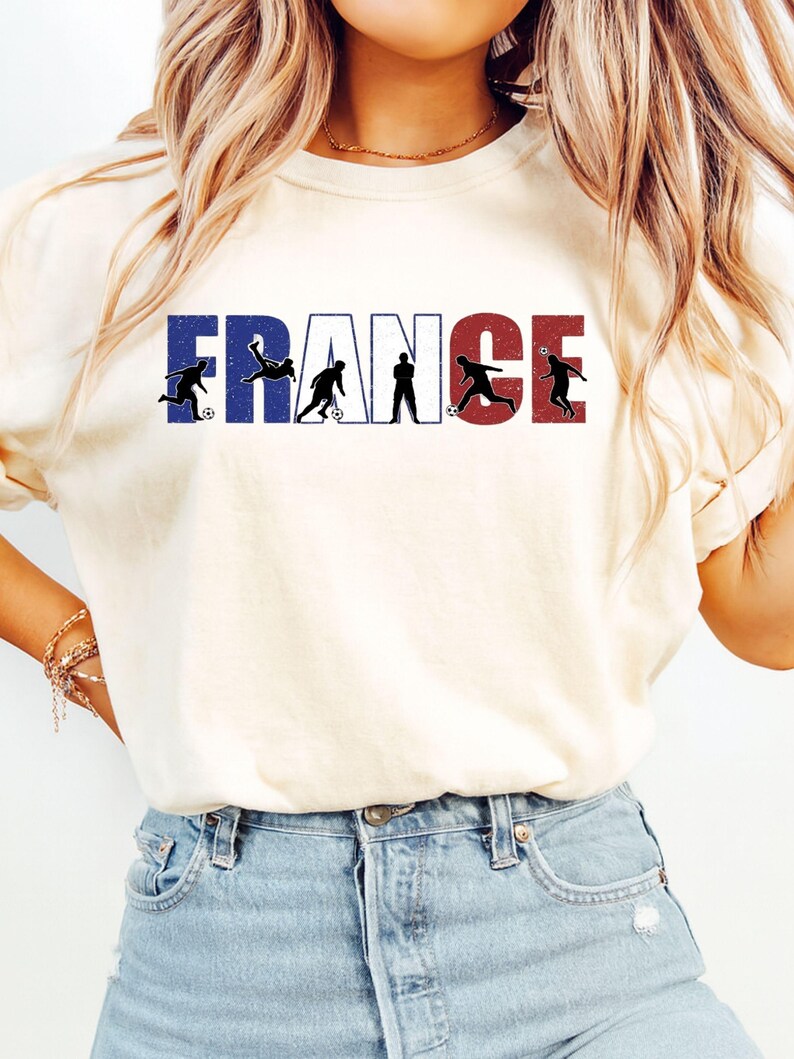May include: Cream-colored t-shirt featuring the word "FRANCE" in blue, white, and red, with silhouettes of soccer players. The shirt has short sleeves and a crew neck.