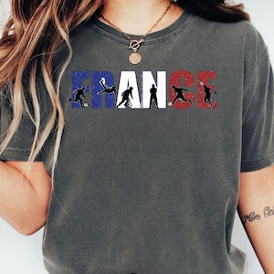May include: Dark gray t-shirt featuring the word "FRANCE" in white, blue, and red, with silhouettes of soccer players incorporated into the letters. The shirt has a crew neck and short sleeves.