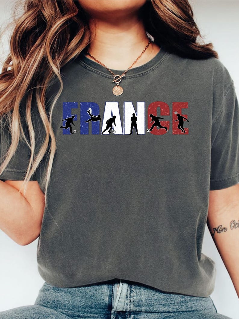 May include: Dark gray t-shirt featuring the word "FRANCE" in white, blue, and red, with silhouettes of soccer players incorporated into the letters. The shirt has a crew neck and short sleeves.