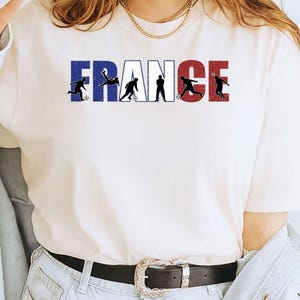 May include: White t-shirt featuring the word "FRANCE" in blue, white, and red, with silhouettes of soccer players incorporated into the letters. The shirt is paired with light-wash jeans and a black belt with a silver buckle.