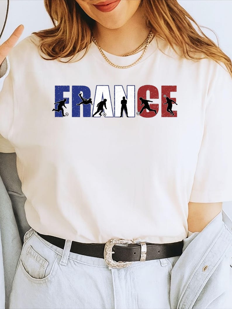 May include: White t-shirt featuring the word "FRANCE" in blue, white, and red, with silhouettes of soccer players incorporated into the letters. The shirt is paired with light-wash jeans and a black belt with a silver buckle.