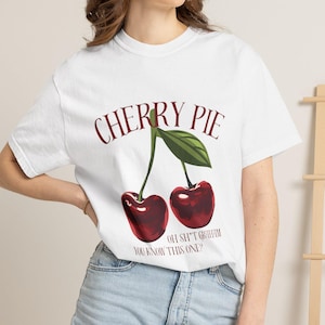 Puede incluir: Una camiseta blanca de manga corta con un gráfico de dos cerezas rojas y una hoja verde. El texto CHERRY PIE está impreso en una fuente arqueada de color rojo oscuro sobre las cerezas, con el texto OH SH*T GROWIN YOU KNOW THIS ONE? debajo.