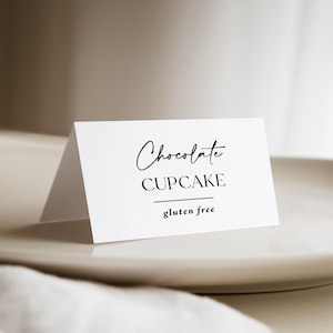 May include: A white, folded card with the words "Chocolate CUPCAKE" in elegant black script. Below, a line and the words "gluten free" are printed. The card is on a white plate, suggesting a dessert or food label.