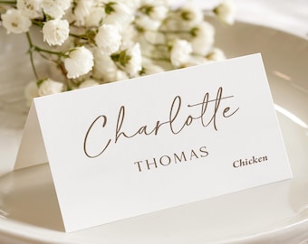May include: A white folded place card featuring the name Charlotte in a brown script font, the surname THOMAS in a brown sans-serif font, and the word Chicken in a smaller brown font, positioned on a plate with white flowers.