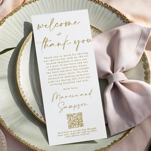 May include: A wedding reception place setting with a white card featuring gold calligraphy that reads "welcome + thank you." The card includes a QR code and a message. A pink napkin is folded on a decorative plate.