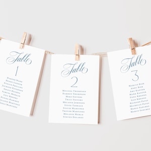 May include: Three white cards with elegant blue text are suspended by wooden clothespins. Each card displays the word "Table" followed by a number and a list of names, ideal for wedding or event seating arrangements.