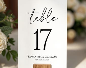 May include: A white rectangular table number card featuring the black text table 17, Samantha & Jackson, and August 27, 2029. The card is held upright by a small, square, light brown wooden block stand with a visible natural wood grain texture.