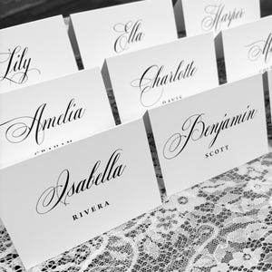 May include: A collection of white folded place cards featuring black printed names in a decorative script font. Each card displays a first name in large cursive lettering with a surname printed in smaller, capitalized sans-serif font below.