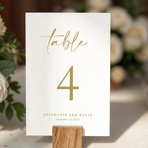 May include: A white table number card with gold script that reads "table" and the number "4". The card also includes the names "Stephanie and Kevin" and the date "September 23, 2029". The card is held in a wooden block.