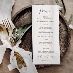 May include: A white rectangular menu card with black and brown text, featuring sections for Salad, Entree, Sides, and Dessert. The menu lists food items and the names Colton & Sampson with the date September 23, 2029, placed on a dark wooden plate.