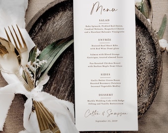 May include: A white rectangular menu card with black and brown text, featuring sections for Salad, Entree, Sides, and Dessert. The menu lists food items and the names Colton & Sampson with the date September 23, 2029, placed on a dark wooden plate.