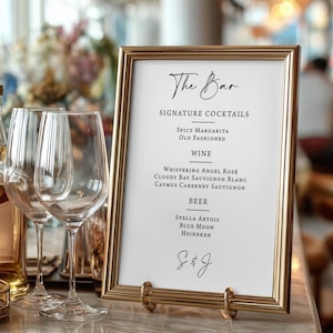May include: A gold framed sign with the words "The Bar" in elegant script. The sign lists signature cocktails, wines, and beers. Two wine glasses are in the foreground, and the background is blurred.