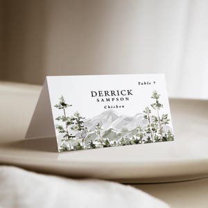 May include: A white folded place card featuring a watercolor illustration of green pine trees and grey mountains. The text reads Table 7, DERRICK SAMPSON, and Chicken in black font. The card is positioned on a smooth, light-colored surface.