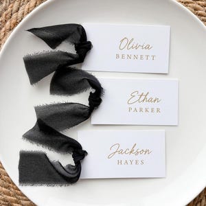 May include: Three rectangular white place cards with gold printed names Olivia Bennett, Ethan Parker, and Jackson Hayes. Each card is attached to a long, frayed-edge black silk ribbon tied in a knot on the left side of the paper card.