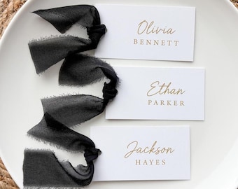 May include: Three rectangular white place cards with gold printed names Olivia Bennett, Ethan Parker, and Jackson Hayes. Each card is attached to a long, frayed-edge black silk ribbon tied in a knot on the left side of the paper card.