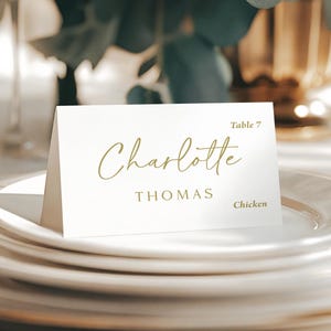May include: A white folded place card with gold text. The name Charlotte is written in a cursive script, followed by the name THOMAS in a sans-serif font. The text Table 7 and Chicken are printed in the upper right and lower right corners.