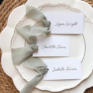 May include: White place cards with names written in black script, tied with sage green ribbons, rest on a white scalloped plate with a gold trim. The plate sits on a woven placemat. Names include Logan Wright, Charlotte Davis, and Isabella Rivera.
