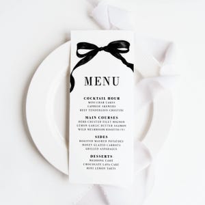 May include: A white menu card with black text and a black ribbon bow. The menu lists cocktail hour, main courses, sides, and desserts. The menu is on a white plate with white fabric in the background.