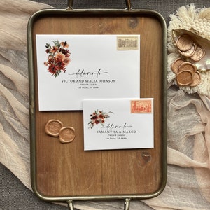 May include: Two white envelopes with floral designs and wax seals. The envelopes are addressed to Victor and Stacia Johnson and Samantha & Marco. The envelopes are on a wooden tray with a brown background.