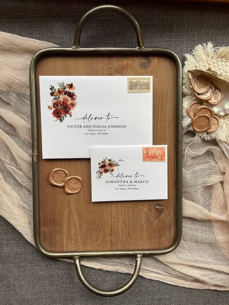 May include: Two white envelopes with floral designs and wax seals. The envelopes are addressed to Victor and Stacia Johnson and Samantha & Marco. The envelopes are on a wooden tray with a brown background.