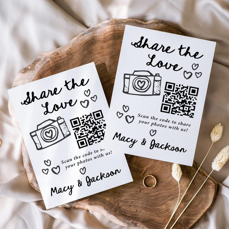 K&ouml;nnte beinhalten: Zwei wei&szlig;e Karten mit dem Text "Share the Love" und einer Kamera-Illustration. Jede Karte hat einen QR-Code und die Namen "Macy & Jackson". Die Karten sind mit Herzsymbolen verziert und liegen auf einer Holzoberfl&auml;che.