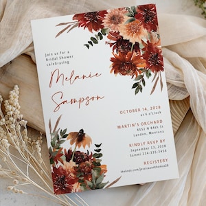 May include: A white invitation card for a bridal shower, featuring floral watercolor artwork in shades of burgundy, orange, and green. The card includes the names "Melanie Sampson" and event details, such as the date, time, and location.