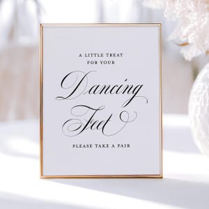 May include: A framed sign with a gold border and white background. The sign reads "A LITTLE TREAT FOR YOUR Dancing Feet PLEASE TAKE A PAIR" in elegant black script. The sign is likely for a wedding or party.