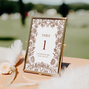 May include: A gold-framed table number card with a floral design. The card displays the number 1 in brown, with the word "TABLE" above it. The names "CATHERINE & JAMES" and the date "AUGUST 19TH, 2028" are also printed on the card.