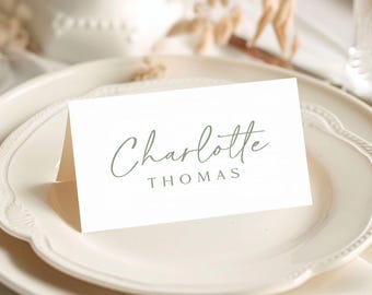 May include: A white folded place card featuring the name Charlotte in a sage green cursive script font, with the surname THOMAS printed below in a simple, uppercase sage green sans-serif typeface, resting on a cream-colored ceramic plate.