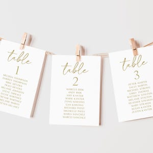 May include: Three white cards with gold text, each labeled "table" followed by a number. The cards are clipped to a string with wooden clothespins. Each card lists names, likely for a seating arrangement at an event.