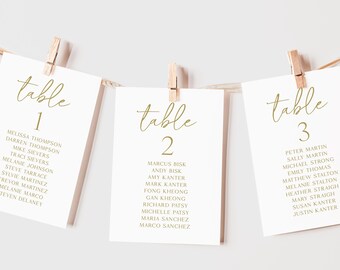 May include: Three white cards with gold text, each labeled "table" followed by a number. The cards are clipped to a string with wooden clothespins. Each card lists names, likely for a seating arrangement at an event.