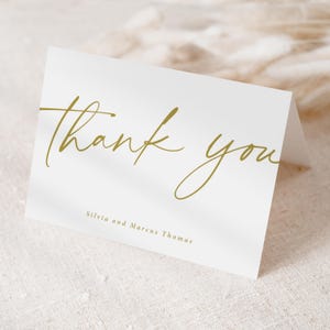 Pu&ograve; includere: Un biglietto bianco piegato con le parole "thank you" in corsivo dorato. I nomi "Silvia and Marcus Thomas" sono stampati in un carattere pi&ugrave; piccolo sotto il testo principale. Il biglietto &egrave; su una superficie strutturata bianco sporco.