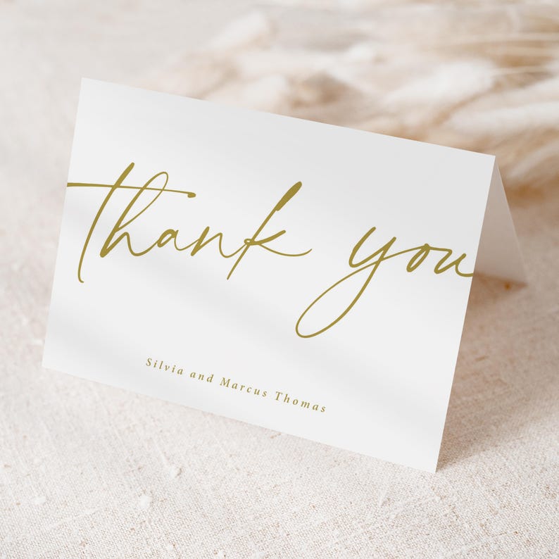 Pu&ograve; includere: Un biglietto bianco piegato con le parole "thank you" in corsivo dorato. I nomi "Silvia and Marcus Thomas" sono stampati in un carattere pi&ugrave; piccolo sotto il testo principale. Il biglietto &egrave; su una superficie strutturata bianco sporco.