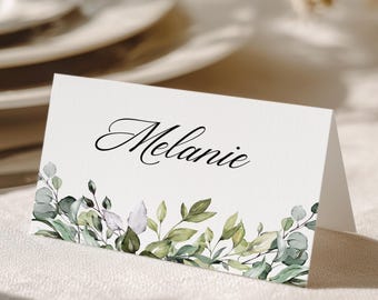 May include: A white folded place card featuring the name Melanie printed in a black cursive script. The bottom edge of the card is decorated with a detailed illustration of green eucalyptus leaves and various botanical foliage.