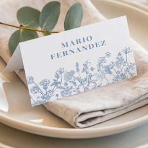 May include: A white place card with the name "MARIO FERNANDEZ" in blue lettering, and a blue floral design at the bottom. The card rests on a folded linen napkin, with a sprig of eucalyptus and a white plate in the background.