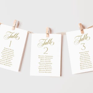 May include: Three white rectangular table seating cards with gold calligraphy text reading Table 1, Table 2, and Table 3, each listing guest names below the number. The cards are suspended from a thin string by small wooden clothespins.
