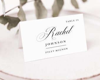 May include: A white folded place card with black text. The card displays the text TABLE 15, the name Rachel Johnson in a script font, and the text FILET MIGNON below a horizontal line. The card is positioned on a light-colored plate.