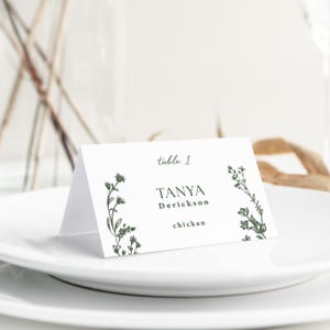 May include: A white folded place card featuring dark green botanical illustrations on the left and right sides. The text reads table 1, TANYA Derickson, and chicken in a mix of serif and script fonts, centered on the front panel of the card.