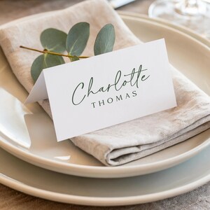 May include: A white folded place card with the name Charlotte Thomas printed in dark green script and block lettering. The card is placed on a folded beige linen napkin, which is topped with a small sprig of green eucalyptus leaves.