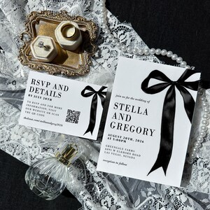 May include: Wedding invitation suite featuring a white invitation with a black bow, and a matching RSVP card. The invitation includes the names Stella and Gregory, and the date August 20th, 2026. A gold tray holds two ring boxes with rings.