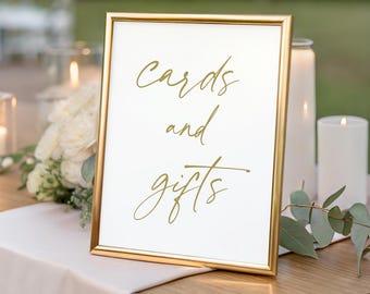 May include: A gold framed sign with the words "cards and gifts" in elegant cursive script. The sign is displayed on a wooden table, with candles and floral arrangements in the background, suggesting a wedding or event.