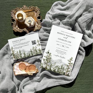 May include: Wedding invitation suite featuring a mountain and pine tree design. Includes invitation, RSVP card, and wax seals. Gold rings in ring boxes are displayed on a decorative tray. The invitation text reads "Monica Walters and Charles Lee".