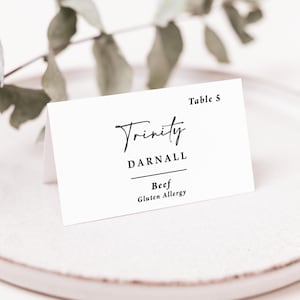 May include: A white folded place card with black text reading Table 5, Trinity, DARNALL, Beef, and Gluten Allergy. The card features a mix of elegant script and clean sans-serif typography, designed for event seating and meal identification.