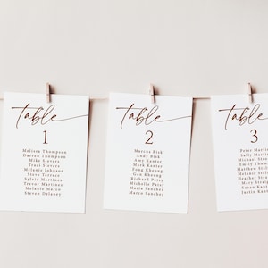 May include: Three white cards with brown text are clipped to a string. Each card reads "Table" in a script font, followed by a number. Below the numbers are guest names. The cards are for a wedding or event.