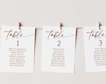 May include: Three white cards with brown text are clipped to a string. Each card reads "Table" in a script font, followed by a number. Below the numbers are guest names. The cards are for a wedding or event.