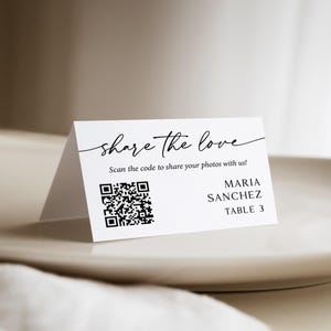 May include: A white folded card with black text and a QR code. The text reads share the love, Scan the code to share your photos with us!, MARIA SANCHEZ, and TABLE 3. The card is designed for event seating or photo sharing instructions.