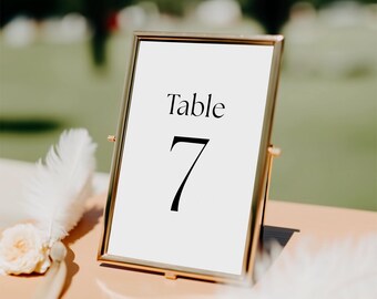 May include: A gold-framed table number sign with the word "Table" and the number "7" in black. The sign is on a peach-colored surface, with white feathers and a flower nearby. The background is blurred.