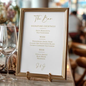 May include: A gold-framed bar menu with the text "The Bar" in elegant script. The menu lists signature cocktails, wines, and beers, including Spicy Margarita, Whispering Angel Rosé, and Stella Artois. The menu is displayed on a marble surface.