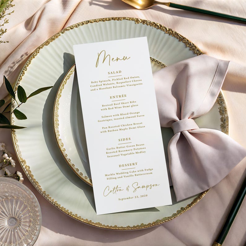 Pu&ograve; includere: Un biglietto del menu di nozze con scritta dorata su sfondo bianco, posto su un piatto decorativo con bordo dorato. Sono visibili anche un tovagliolo rosa piegato e un cucchiaio d'oro, che suggeriscono un ambiente formale.