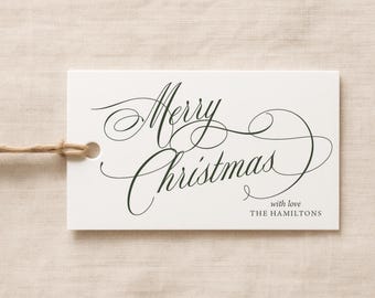 May include: A rectangular white gift tag with a hole on the left side threaded with brown twine. The tag features dark green cursive text that reads Merry Christmas, with the smaller text below reading with love THE HAMILTONS.